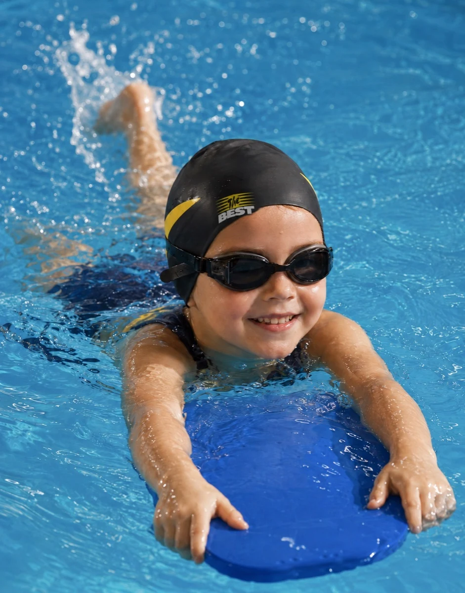 Criança sorridente com touca The Best preta e amarela nadando com prancha na piscina aquecida